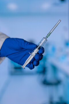 Blue-gloved hand holds a syringe. Clear liquid fills the syringe tube. Blurred lab equipment forms the background. Medical precision is evident. Ideal for healthcare or research contexts