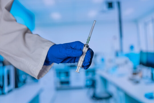 Close-up of a gloved hand holding a cylindrical metal object. The background shows a blue-tinted laboratory with blurred equipment. The person wears a white lab coat and blue gloves