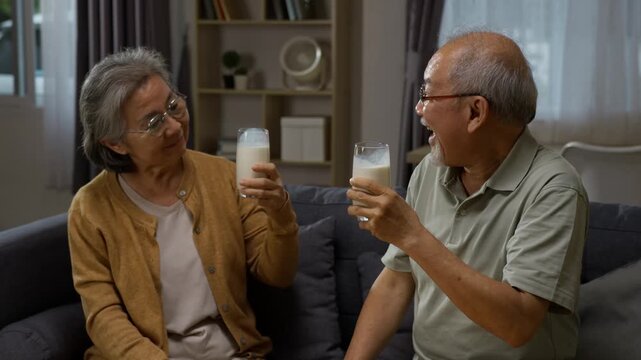 Happy Asian elderly husband and wife toasting and drinking milk together on the sofa. Promoting healthy diet and strong bones for older adults.