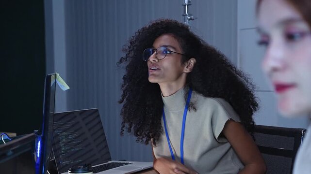 Focused female programmer resting hand on chin at office workstation with computer screen and laptop displaying code scripts during software development project session