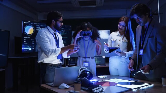 Software development team testing virtual reality headset and robotic prosthetic hardware prototype near screen monitor displaying data graphics during office technology workshop