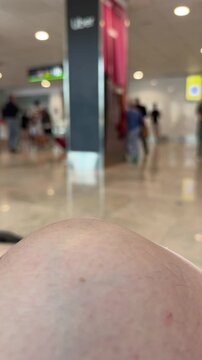 First-person perspective of someone sitting and waiting in a busy concourse. The background shows many people walking in a large, bright hall