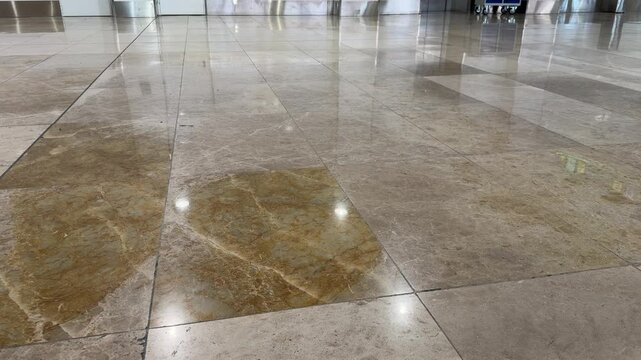Low angle view of a polished marble floor in a large hall. Reflections of walking people and their belongings are visible on the glossy tiles