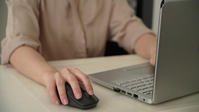 Close up of a woman using a wireless computer mouse beside a laptop on an office desk, with her other hand reaching toward the keyboard. Everyday computer work and office technology.
