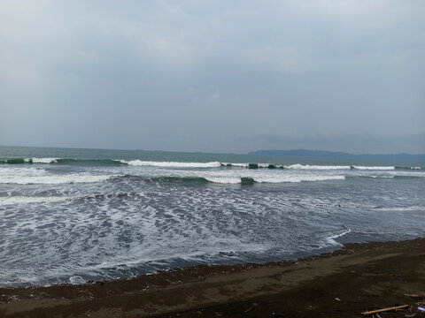 A wide-angle landscape photograph capturing the dynamic energy of the ocean on an overcast day. White-capped waves roll rhythmically onto a dark, volcanic sand beach, creating intricate foam patterns 