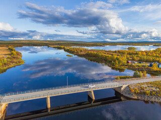 Aerial view of a white car crossing a modern bridge over a mirror-like lake reflecting a dramatic cloudy sky in arctic Lapland, Sweden, with golden autumn foliage © John N