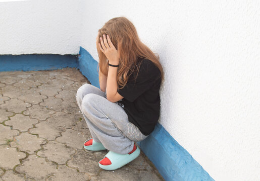 A teenage girl is sitting on her heels near a white wall, clutching her head in her hands; the concept conveys feelings of resentment, loneliness,