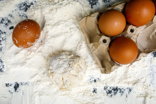 Brown eggs and an egg carton resting on a pile of white flour with a small ball of dough on a dark background, high quality photo
