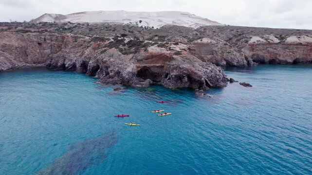 Wide drone shot of colorful kayaks paddling across deep blue sea waters near the rugged cliffs of Tsigrado Beach on Milos Island, Greece