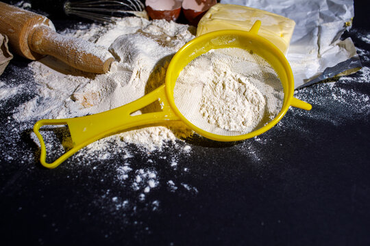 Yellow sieve filled with white flour resting on a dark table with a rolling pin and paper in the background
