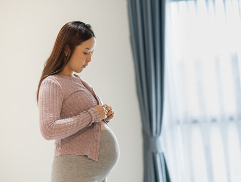 Pregnant woman looking at her baby bump while standing indoors near a window.
