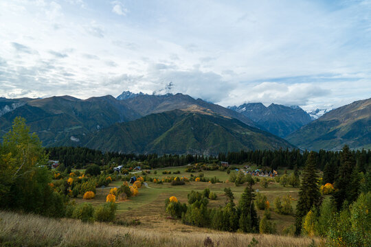Landscape photograph of mountain village with autumn trees and majestic snowy peak under cloudy sky in Svaneti Georgia.