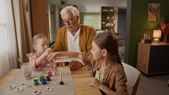 Senior Caucasian man helping two young Caucasian children painting wooden craft model at home. Ideal for retirement lifestyle and intergenerational family campaigns