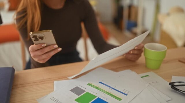 Woman reading a document while holding a smartphone, hands visible over papers on desk in a studio; focus.