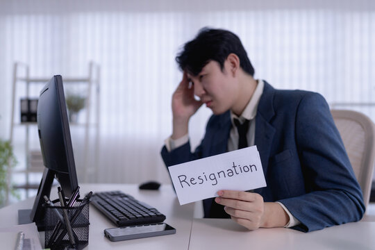 Stressed Asian businessman holding resignation letter at office desk while touching head in modern workspace