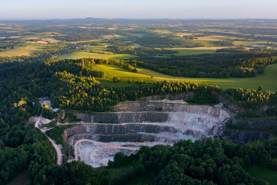 Aerial view of a large open pit stone quarry mine with terraced rock layers surrounded by green landscape during the sunset