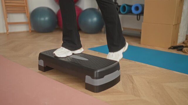 Woman in white trainers stepping onto black aerobic step platform with feet balancing near blue mat and exercise balls in studio; focus balance strength.
