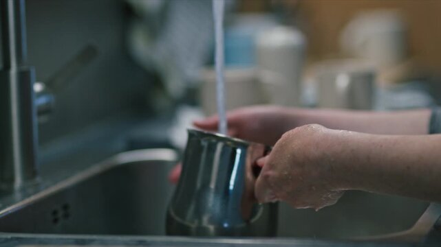 Hands rinse a stainless steel pitcher under a running faucet in a home kitchen sink, capturing the everyday chore of dishwashing, cleaning and domestic routine with natural, soft lighting. 4K