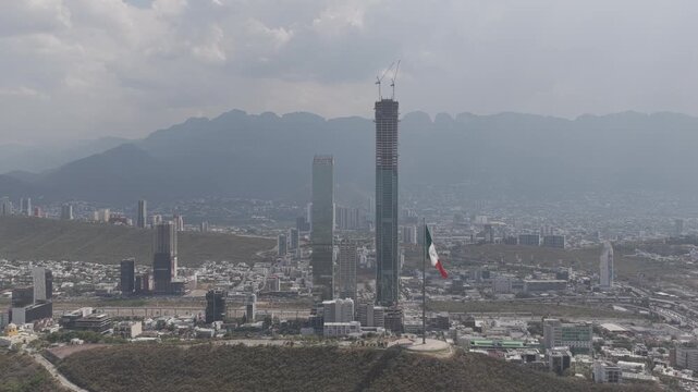 Aerial panoramic view, drone hovering in front of the Mexican Flag at Obispado with Torre Rise and the San Pedro Skyline in the background. Shot in D-Log M