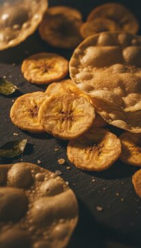 Crispy banana chips and papadum with salt flakes and curry leaves on a dark textured surface, close-up with warm lighting