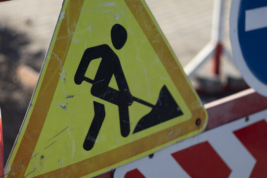 A warning sign indicates road work taking place on a street. The sign shows a person using a shovel. Barriers and traffic signs are also visible around the area