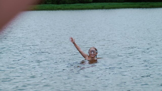 In the foreground, a mother's hand waves to her young son as he swims in a lake wearing a diving mask. The scene captures family connection, childhood, and summer recreation.