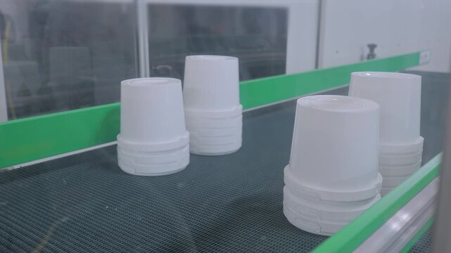 Stacks of white plastic yogurt containers are moving on conveyor belt of plastic injection molding machine with pick and place robotic arm manipulator at exhibition, factory - close up view