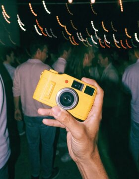 A spontaneous capture of a hand gripping a disposable film camera surrounded by a lively crowd creating a sense of movement and genuine ambiance