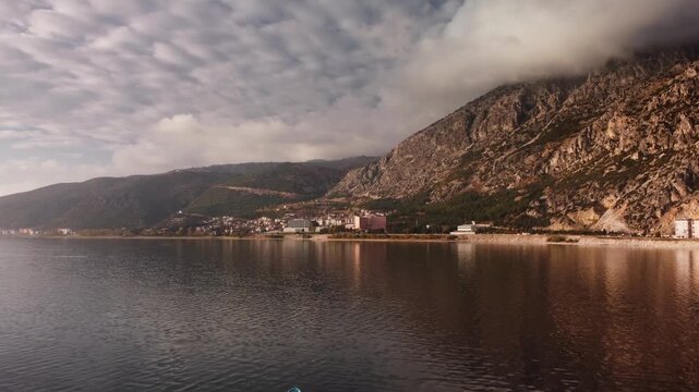 Serene mountain backdrop overlooking small town with waterfront homes and peaceful maritime ambiance. Cozy village and rocky shoreline along the sea at dusk. Narrow promenade and terraced roofs. 