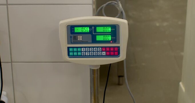 close-up view of digital weighing scale on stainless post in tiled bakery workroom, green lcd readout and keypad visible, cables and textured surfaces softly, with