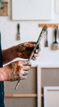 Paint stained hands hold weathered brushes gathered from studio wall rack