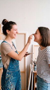 Playful artists laugh as one dabs paint on friend's nose in art studio
