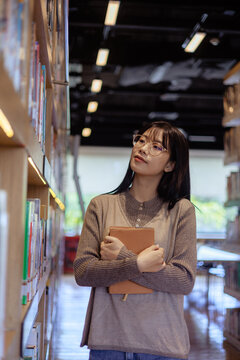 Young Asian Woman with Glasses Holding Book in Library, Freedom to Learn and Explore Knowledge