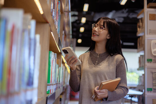 Young Asian Woman Browsing Books in Library While Holding Smartphone , Freedom to Learn and Explore Knowledge