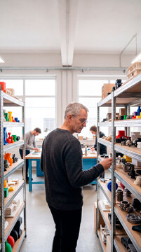 Mature designer examines shelf of colorful 3D printed models in workshop