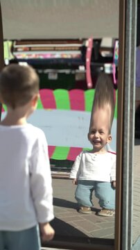 Happy child has fun looking at his distorted reflection in a curved mirror, smiling and laughing at an amusement park on a sunny summer day.