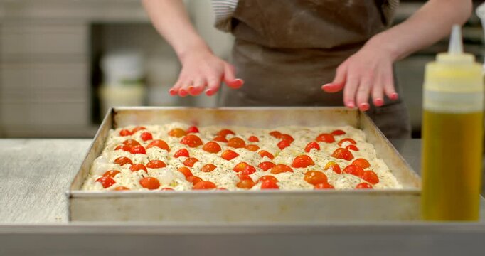 baker presses tomato-studded focaccia dough in rectangular metal pan on stainless countertop, then seasons and oils surface using squeeze bottle and shaker, with