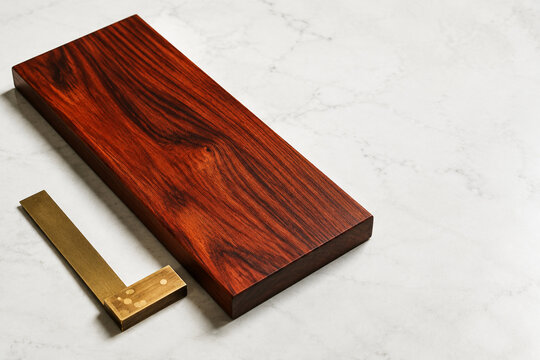 Polished padauk wood slab with brass carpenter's square, bright studio surface, wide crop