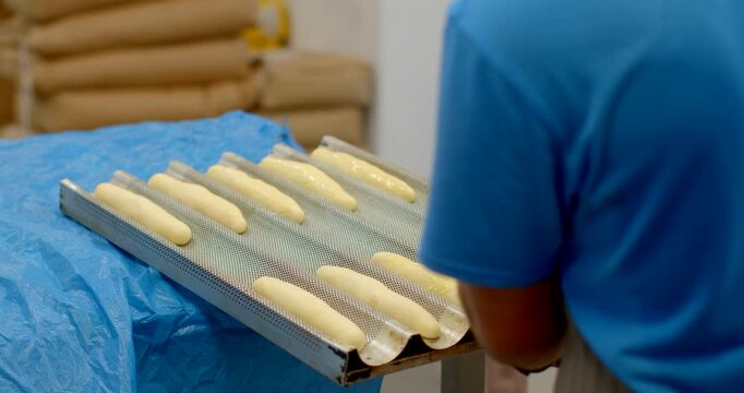 worker in blue shirt applies spray glaze to aligned unbaked baguette-shaped dough in perforated metal tray on prep table, scene shows routine handling and, with