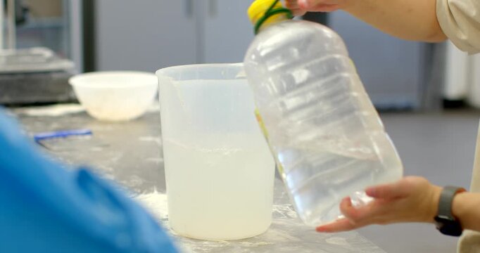 close-up of hands and forearms pouring clear liquid from large plastic bottle into translucent measuring jug on stainless workbench in commercial kitchen or bakery
