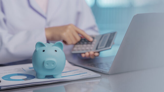 Doctor using a calculator for financial calculation with a blue piggy bank and report on desk. Healthcare professional managing budget, insurance, savings and investment planning.