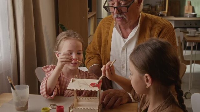 Senior Caucasian grandfather helping two granddaughters paint wooden model at home. Clip ideal for intergenerational bonding, parenting, retirement lifestyle campaigns