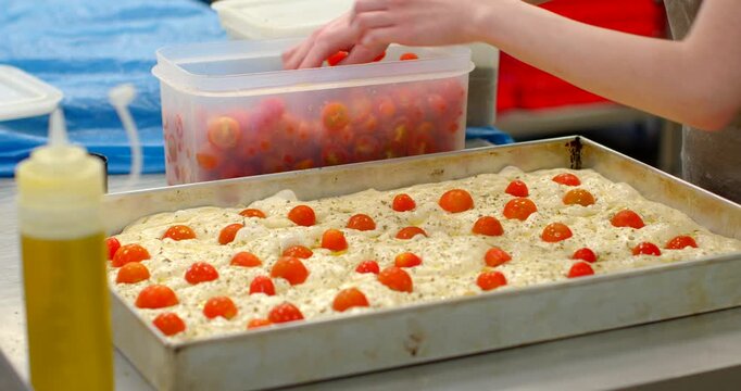 hands of bakery worker carefully place halved cherry tomatoes into dimples of focaccia dough inside large rectangular metal pan, showing assembly and garnish stage
