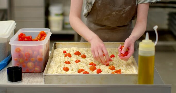 close-up motion of hands arranging halved cherry tomatoes across dimpled focaccia dough in rectangular metal tray on stainless steel prep table inside professional