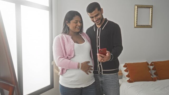 Pregnant woman touching belly while partner holds smartphone for a selfie in a bedroom inside a building, smiling and leaning close; expectation intimacy joy.