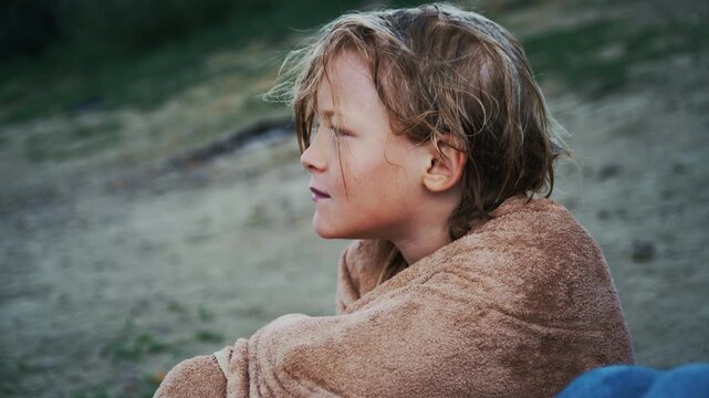 Young boy with wet blond hair shivers while wrapped in a brown towel after swimming in a lake. The footage represents childhood, leisure, outdoor activities, and a chilly moment after water recreation