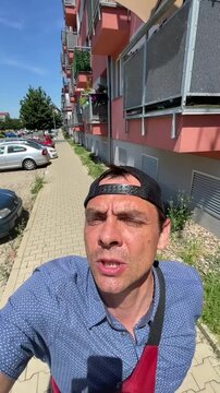 adult man wearing backward baseball cap and blue patterned shirt holds camera for close-up selfie while walking along paved sidewalk beside parked cars and red