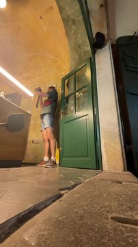 young person stands inside rustic interior holding bottle with both hands at chest level near green half-glass door, low-angle viewpoint from cracked tiled floor