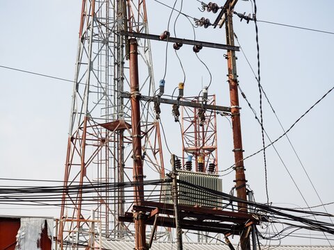 Electrical distribution transformer mounted on utility poles with complex overhead cables and a telecommunication tower background.