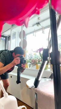 photographer crouches low while photographing tabletop subject, then checks image on camera, scene set in bright window-lined studio or cafe with windowsill potted